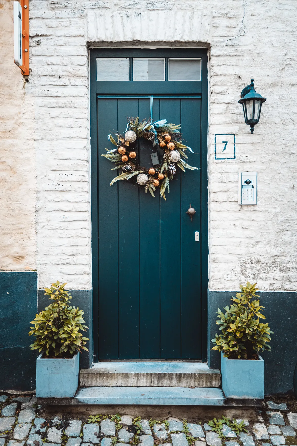 A festive green door with a Christmas wreath, lantern, and potted plants on a cobblestone entry.