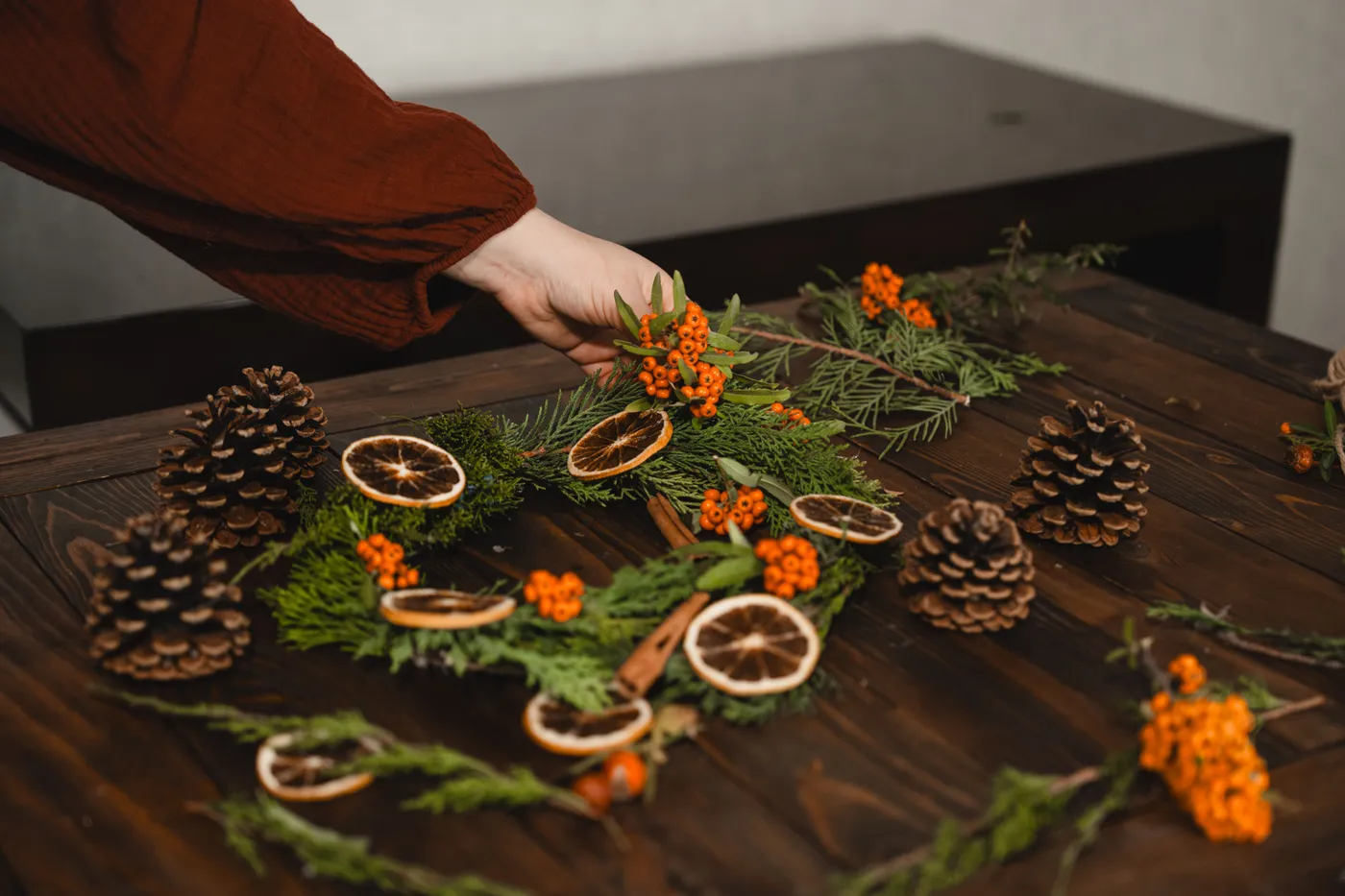 A person arranging a Christmas wreath with pinecones and dried oranges on a wooden table.