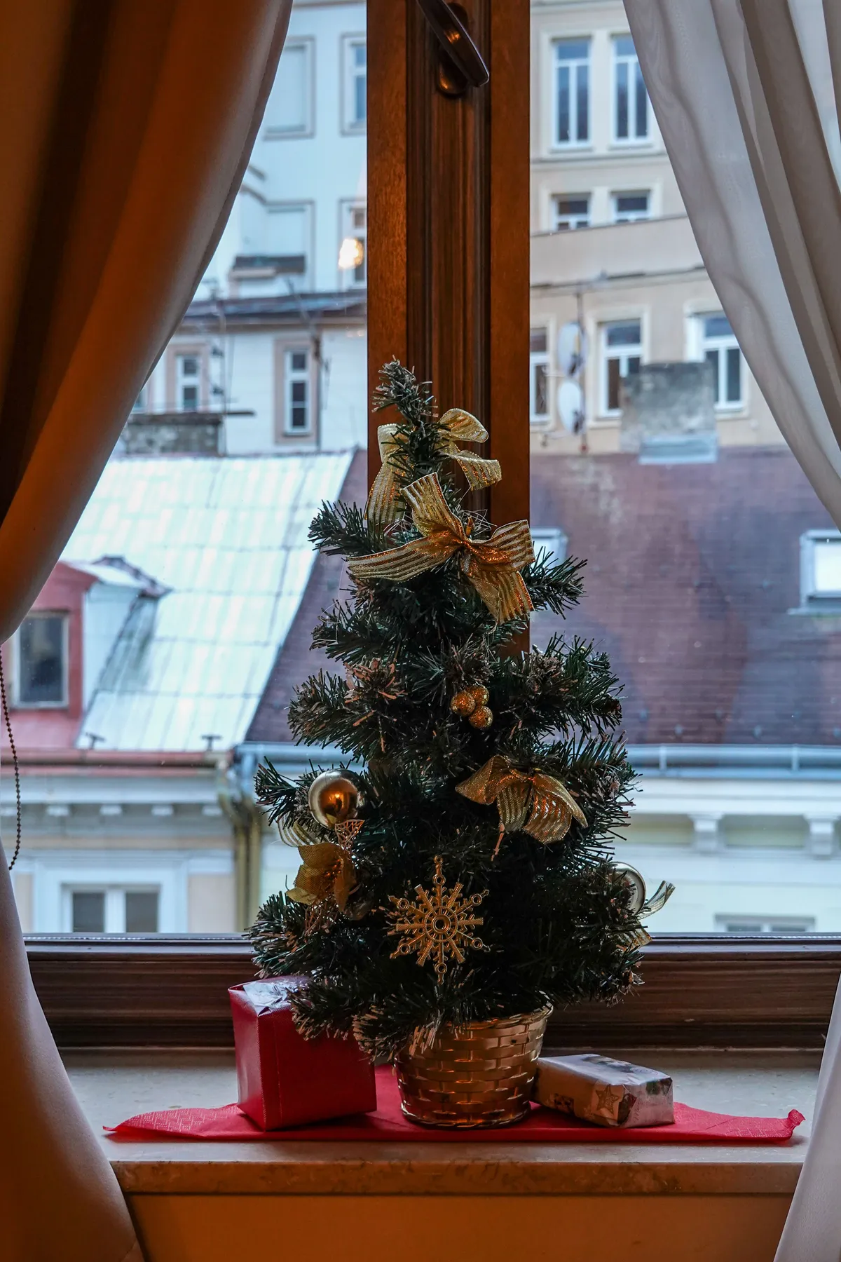 A small Christmas tree with ornaments and gifts on a windowsill overlooking rooftops.