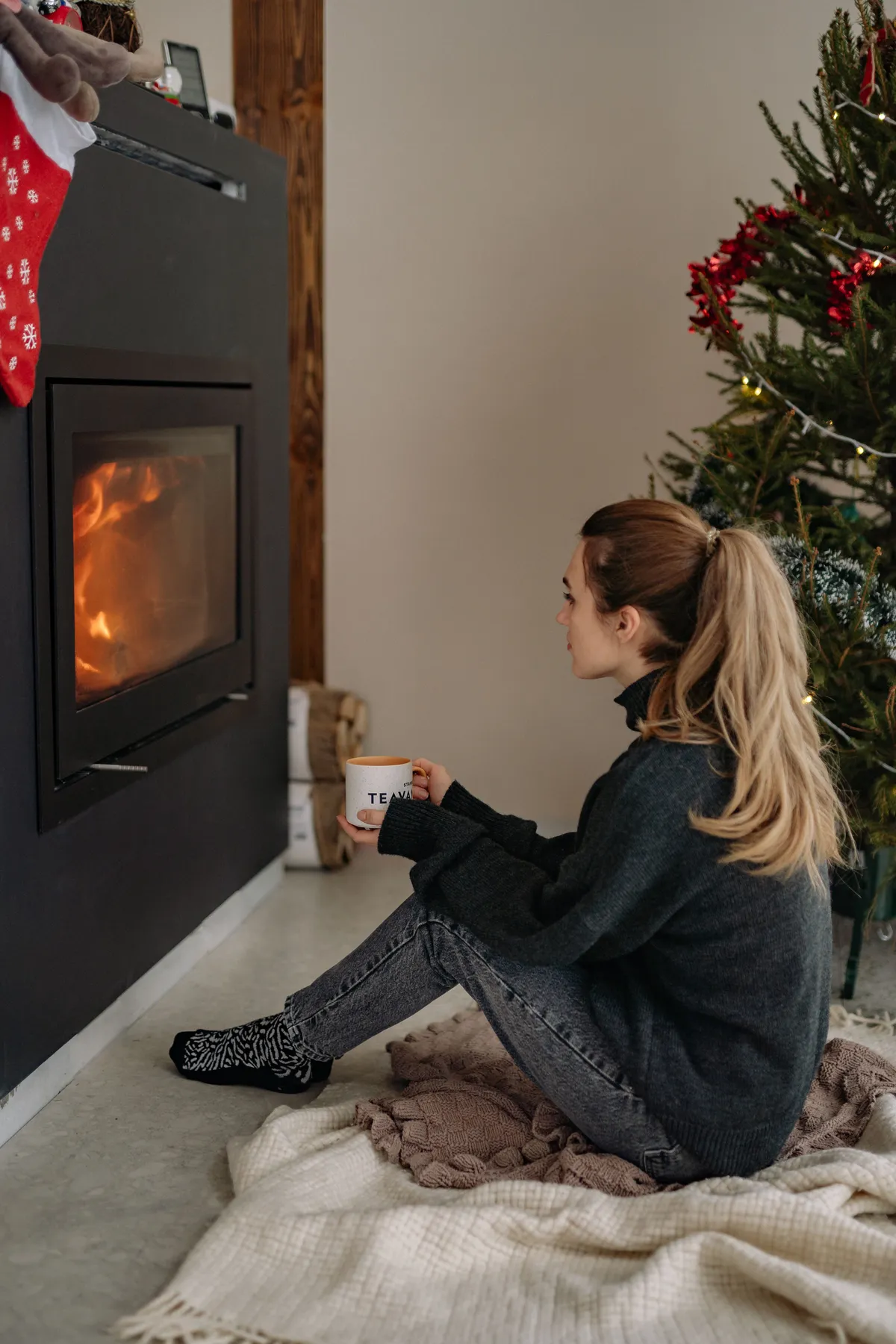 A woman enjoys a warm drink by the fireplace, next to a decorated Christmas tree inside a cozy room.