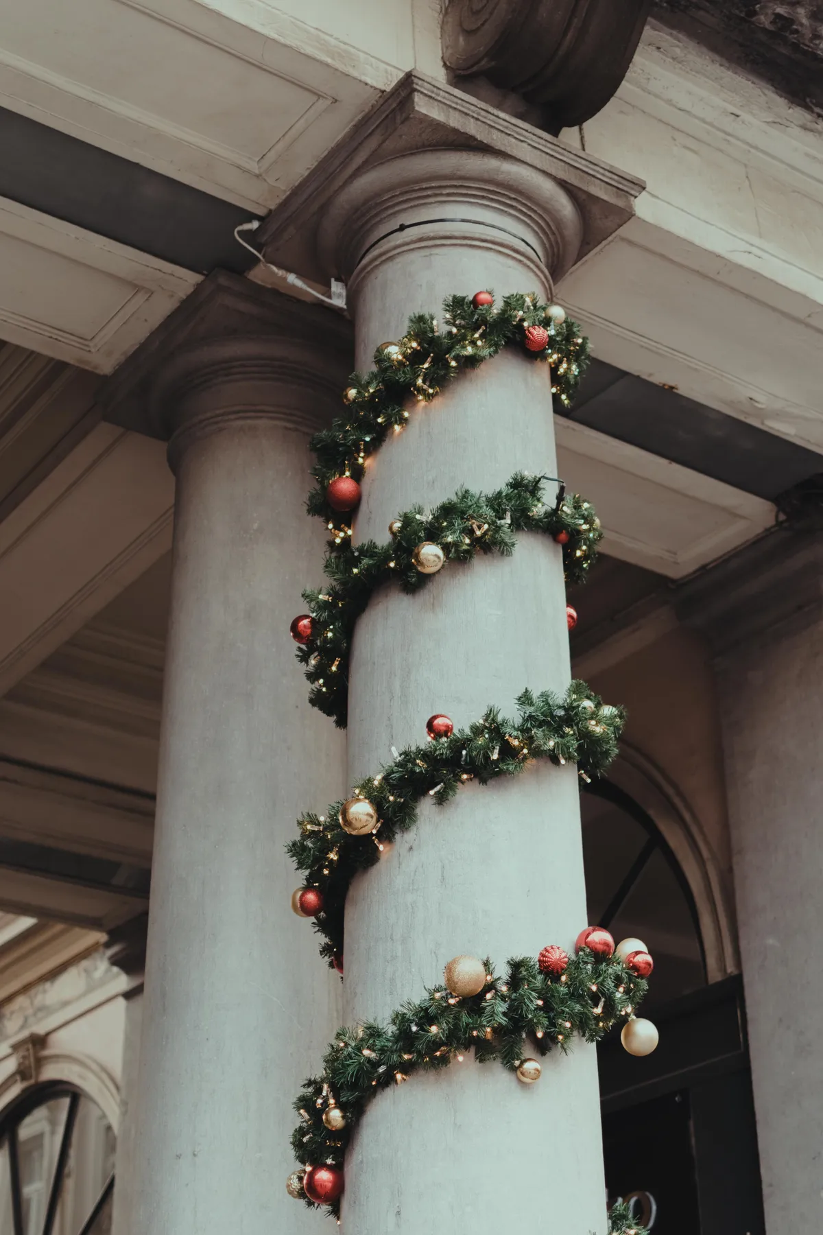 Column wrapped with festive Christmas garland and ornaments, outdoors in a city setting.