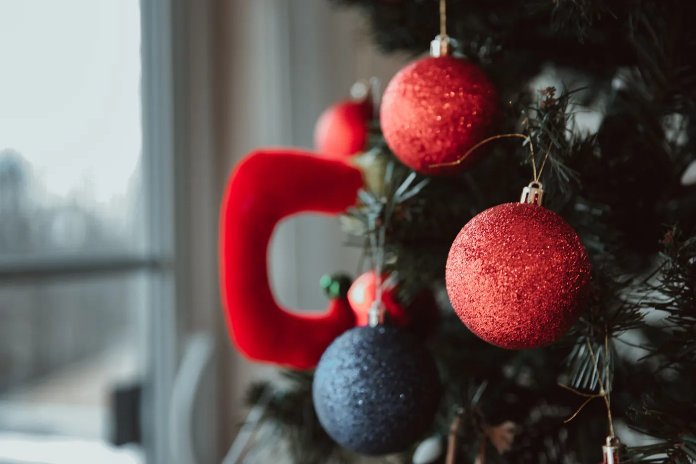 Red and blue decorative baubles hanging on branches of Christmas tree near window on blurred background during holiday preparation in apartment