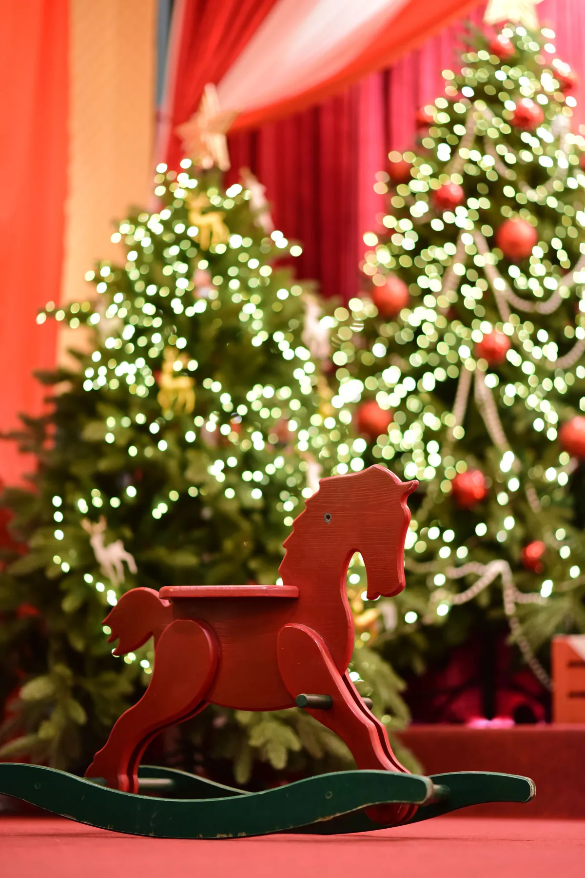 Red wooden rocking horse in front of beautifully decorated Christmas trees with lights.