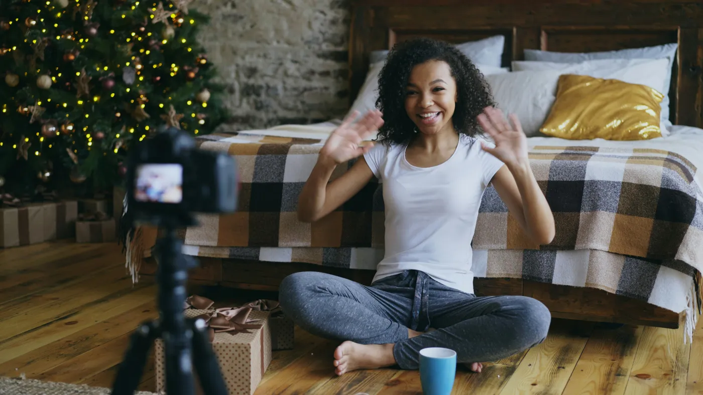 Young cheerful mixed race girl sitting near bed recording video blog about packing christmas gift box with dslr camera at home